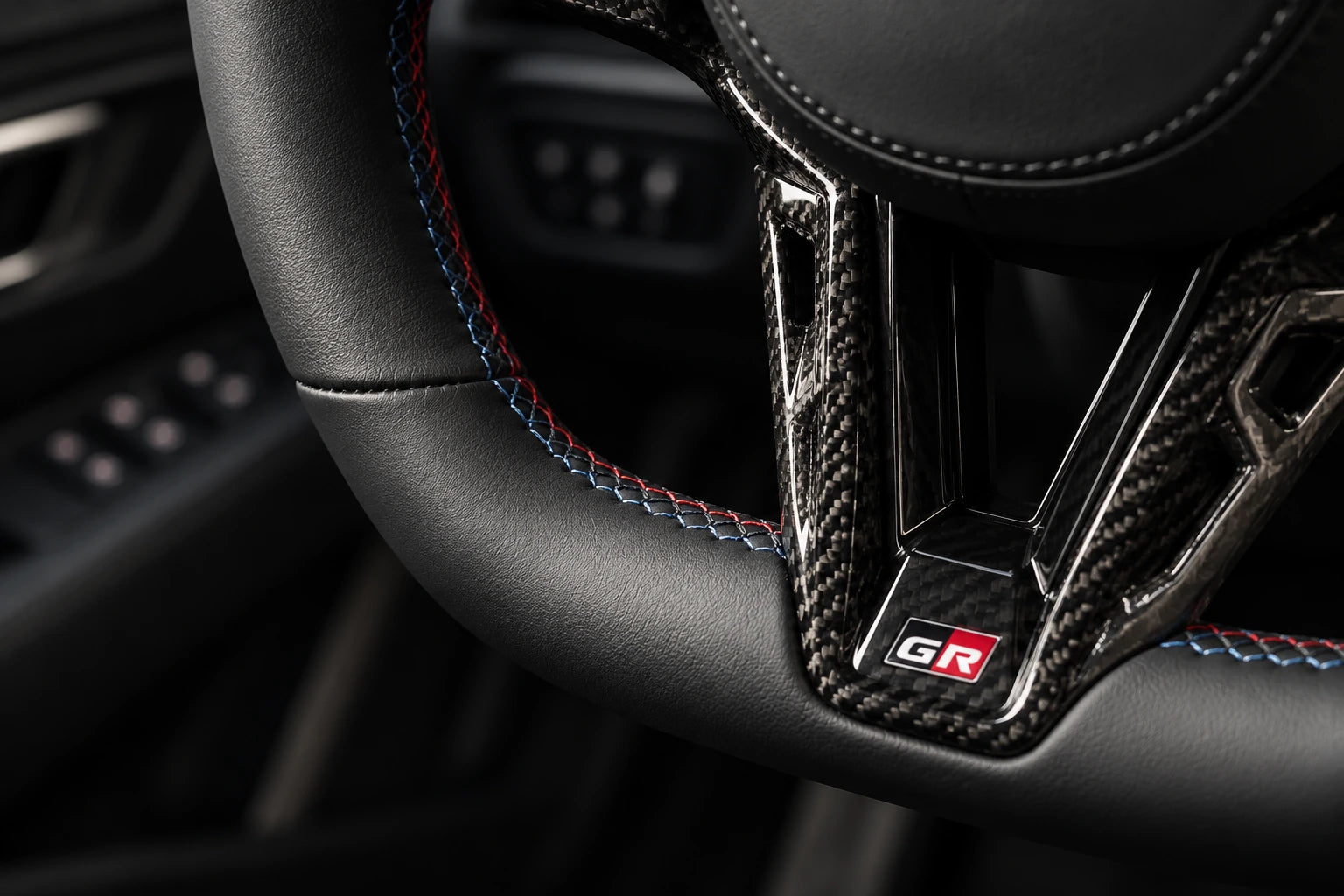 Close-up illustration of black leather, tri-color stitching, carbon fiber and the GR badge on the steering wheel.